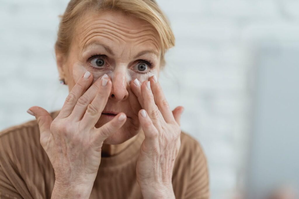 Crop focused mature female applying moisturizing nourishing eye patches and looking away in light bathroom